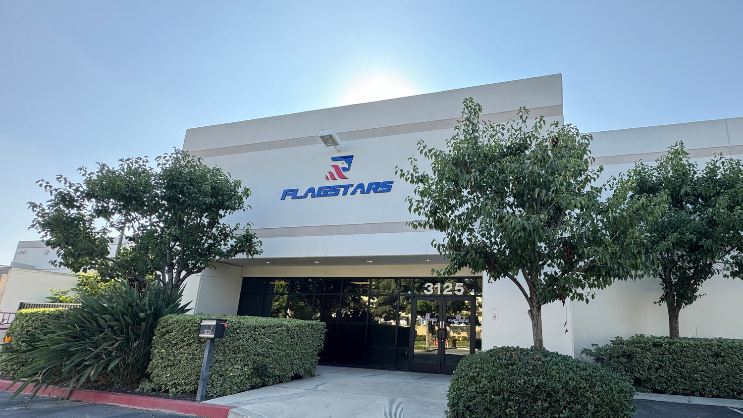 A single-story commercial building with the "Flagstar" logo above the entrance is located at 3125. Trees and shrubs surround the front, and a white car is parked to the right. Known as a reputable manufacturer, this site prides itself on offering high-quality, hand-made products.