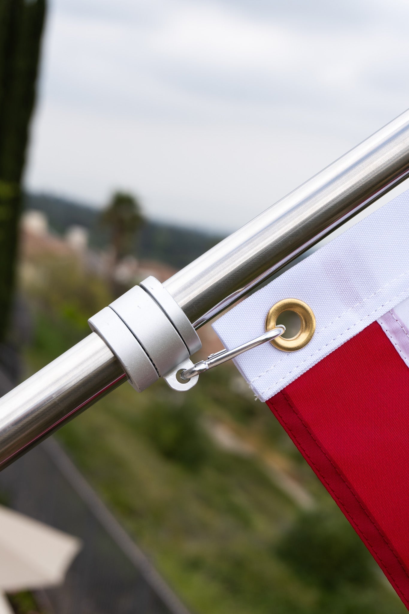 Close-up of an American flagpole with a secured metal clip attached to the grommet of a red and white flag. The background shows an out-of-focus natural landscape, highlighting the long-lasting quality crafted by our manufacturer.