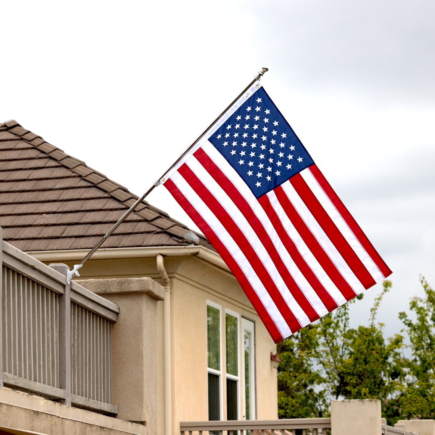An American flag, made in the USA, is mounted on the side of a house with tan siding and a tiled roof, against a backdrop of overcast sky and greenery.