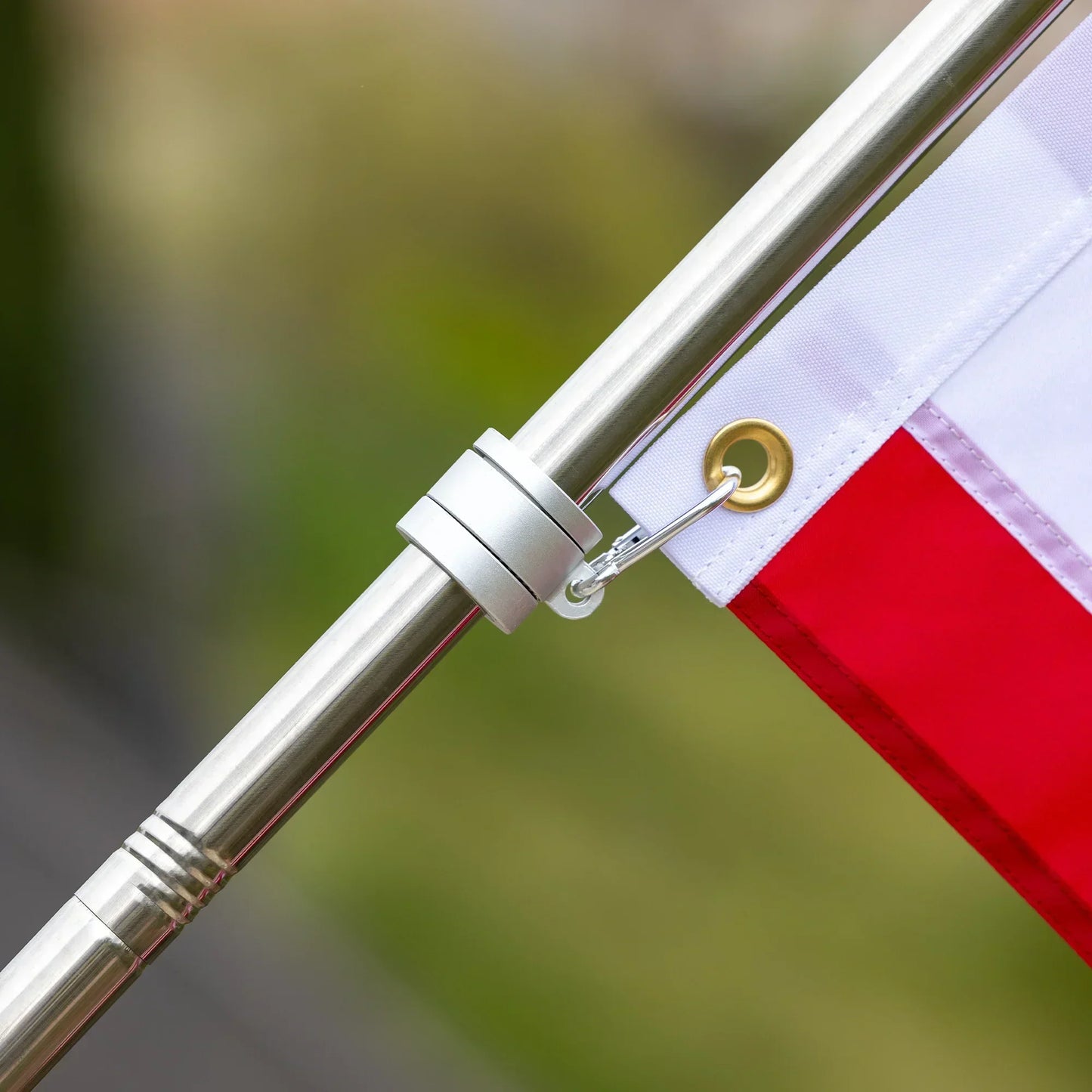 A close-up of a silver, water-resistant flagpole with a metal ring attaching the Poly USA Flag - 100% Made in USA by Flag Stars Inc via its grommet. The background is blurred with green and gray tones.