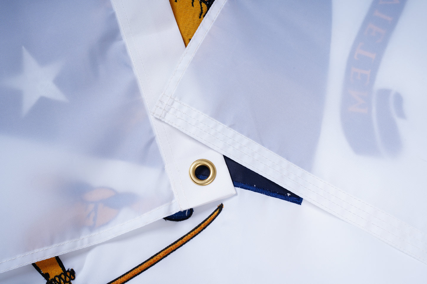 Close-up of the Massachusetts State Flag by Flag Stars Inc—100% Made in USA—showing white fabric with stitched edges, brass grommet, and outdoor durability. Navy blue, gold, and a white star from the printed design are partially visible.