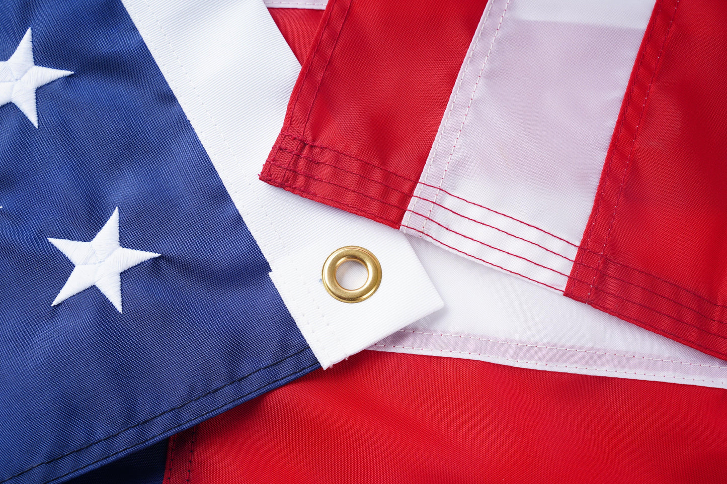 Close-up of folded sections of the Flag Stars Inc Poly USA Flag—100% Made in USA—showing the blue field with white stars, red and white stripes, precise stitching, raised embroidery, and a metal grommet.