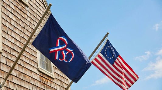 Official America 250 flag and Betsy Ross 13-star flag flying side by side on house-mount poles for the 2026 Semiquincentennial