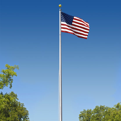 The 6-Section 25FT Aluminum Flagpole by Flag Stars Inc displays the United States flag waving against a clear blue sky, with green trees visible at the bottom edges of the image.