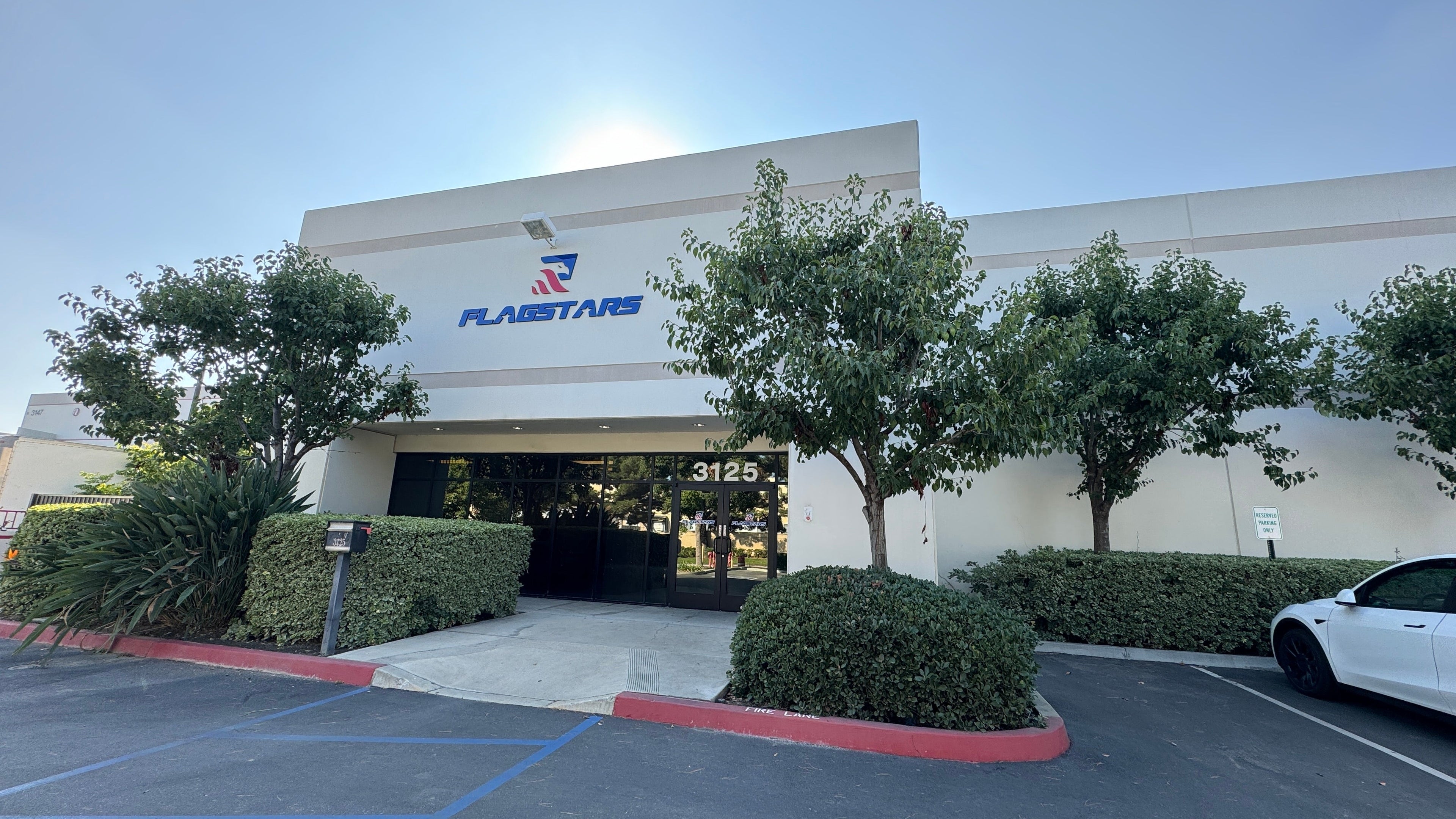 A single-story commercial building with the "Flagstar" logo above the entrance is located at 3125. Trees and shrubs surround the front, and a white car is parked to the right. Known as a reputable manufacturer, this site prides itself on offering high-quality, hand-made products.
