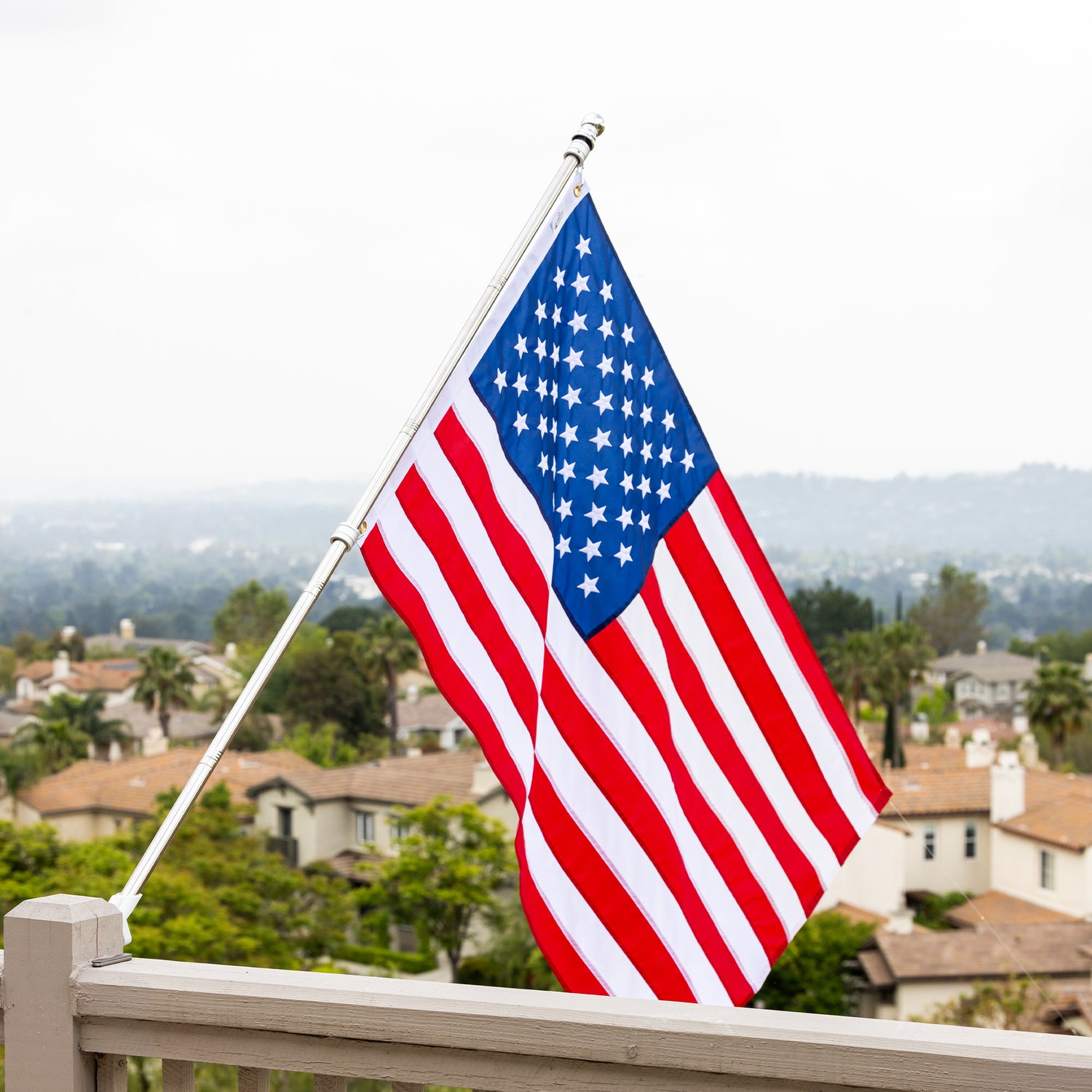 An American flag, hand made and long lasting, is mounted on a pole attached to a wooden railing, with a suburban landscape and trees visible in the background.
