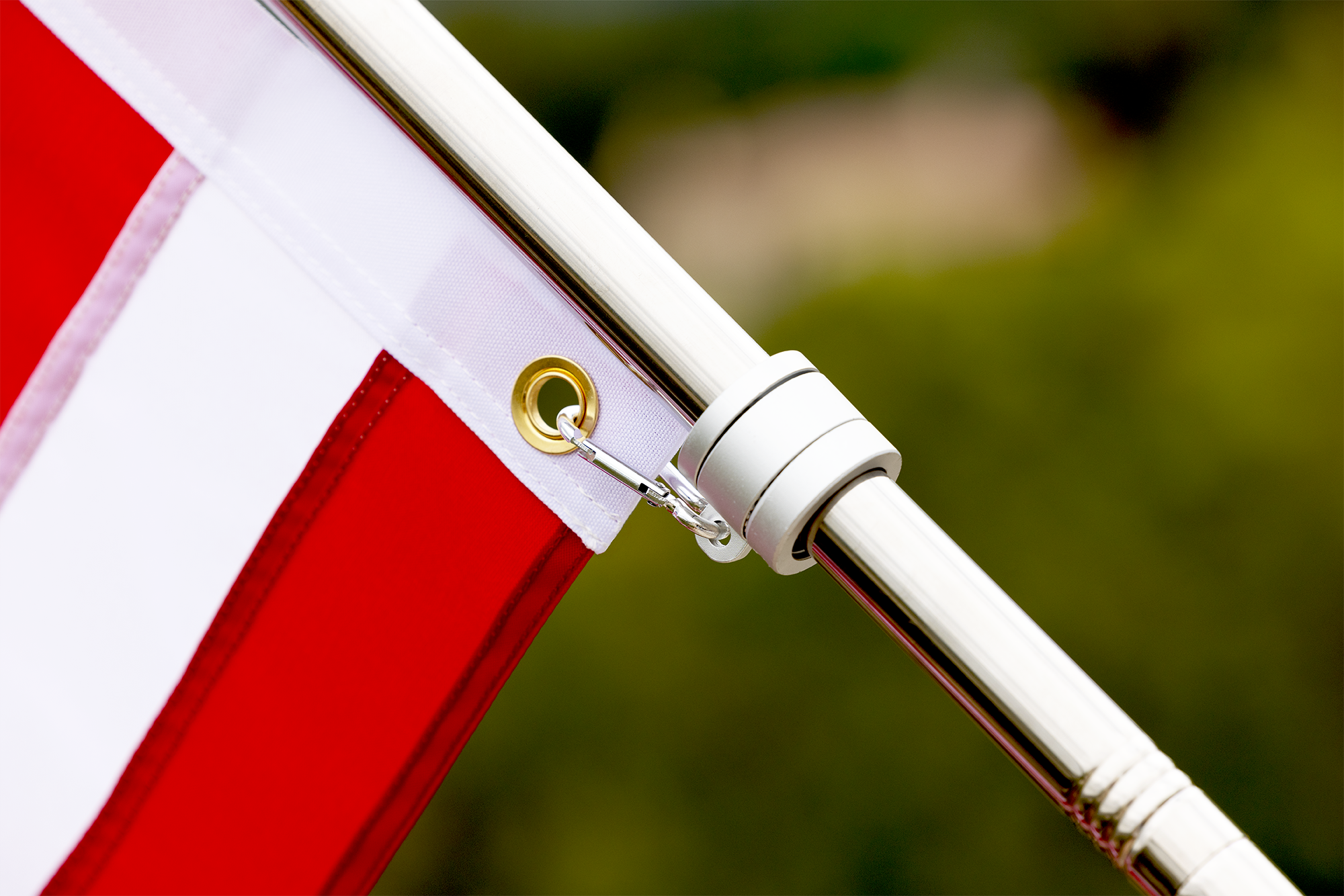 Close-up of a red and white striped flag attached to a pole with a metal grommet and clip, pre-assembled by the manufacturer, with a blurred green background.