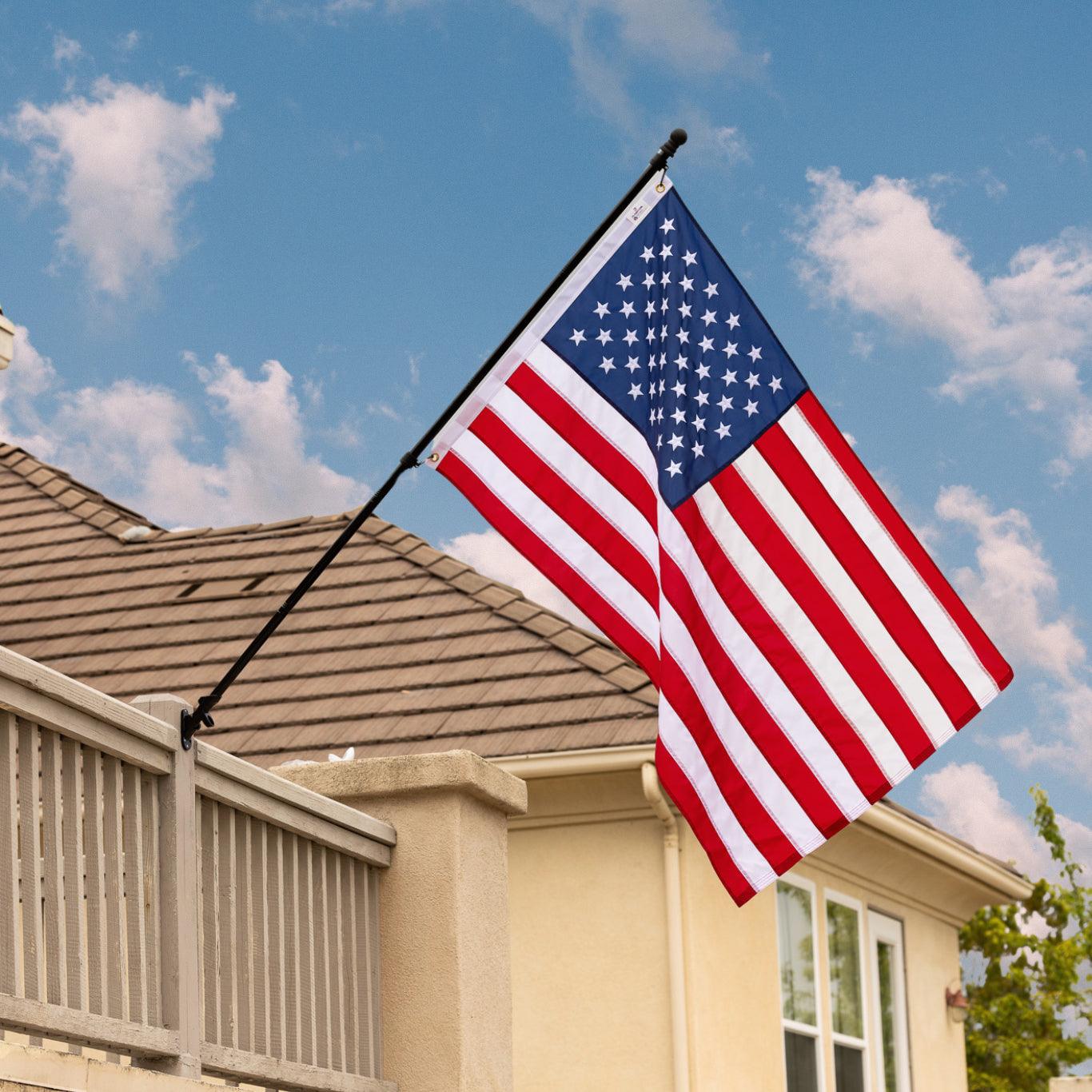 A U.S. flag from the "AMERICAN FLAG SET - COMPLETE BUNDLE" by The FlagStars is mounted on an adjustable aluminum flagpole, attached to the balcony of a house with a tiled roof and beige exterior, under a blue sky with scattered clouds.