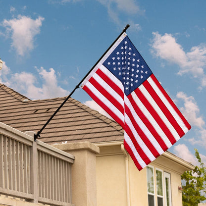 An American flag, easily installed with The FlagStars 6 FT ALUMINUM FLAG POLE WITH TANGLE FREE SPINNERS, is displayed on a pole mounted to the side of a beige house with a tiled roof, set against a background of a blue sky with some clouds.
