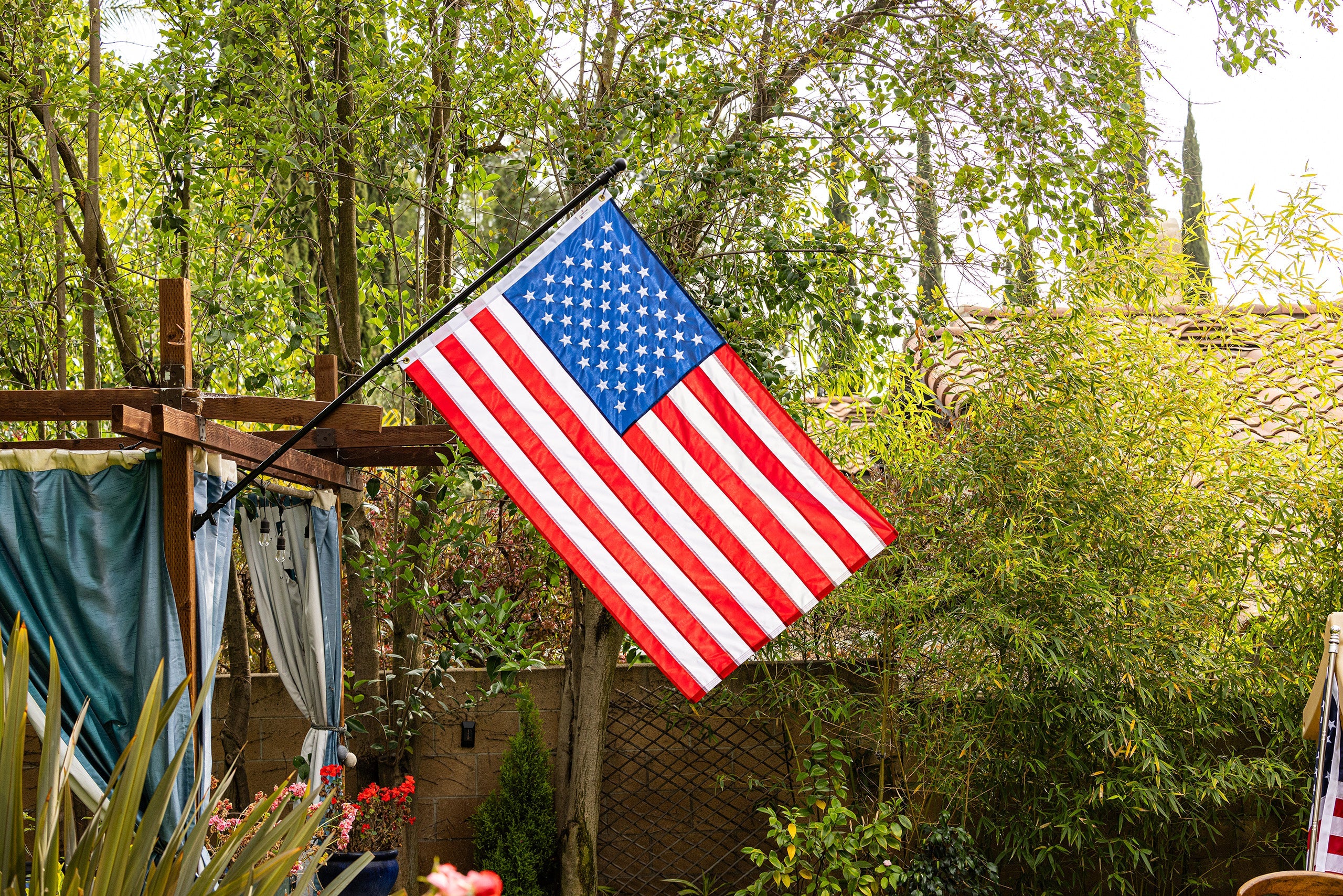An American flag, made in the USA, is displayed on a pole in a lush, green backyard with trees and plants surrounding it.