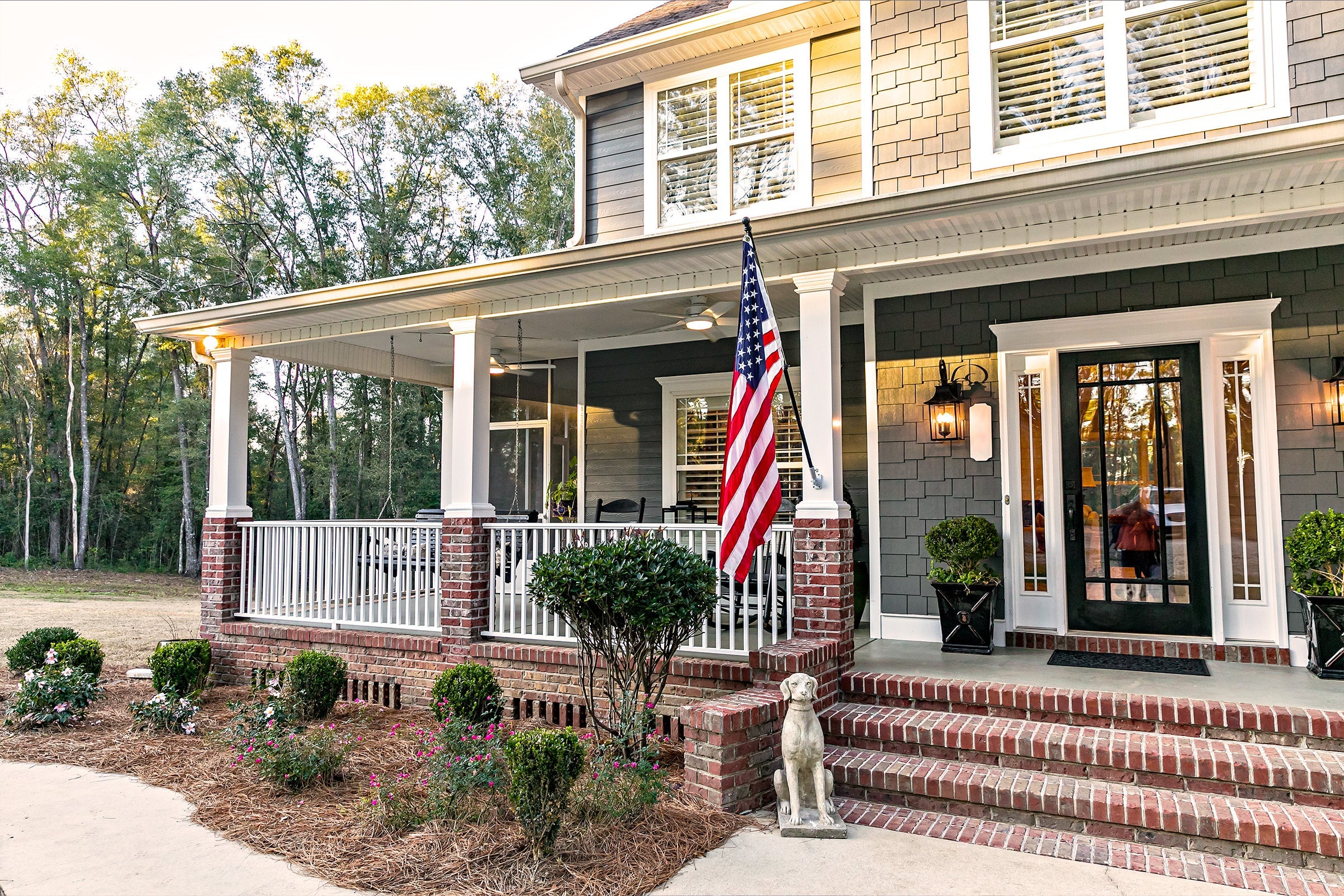 Two-story house with a large front porch, American flag, and brick steps. Hand-made bushes and flowers line the walkway. Forested area in the background.