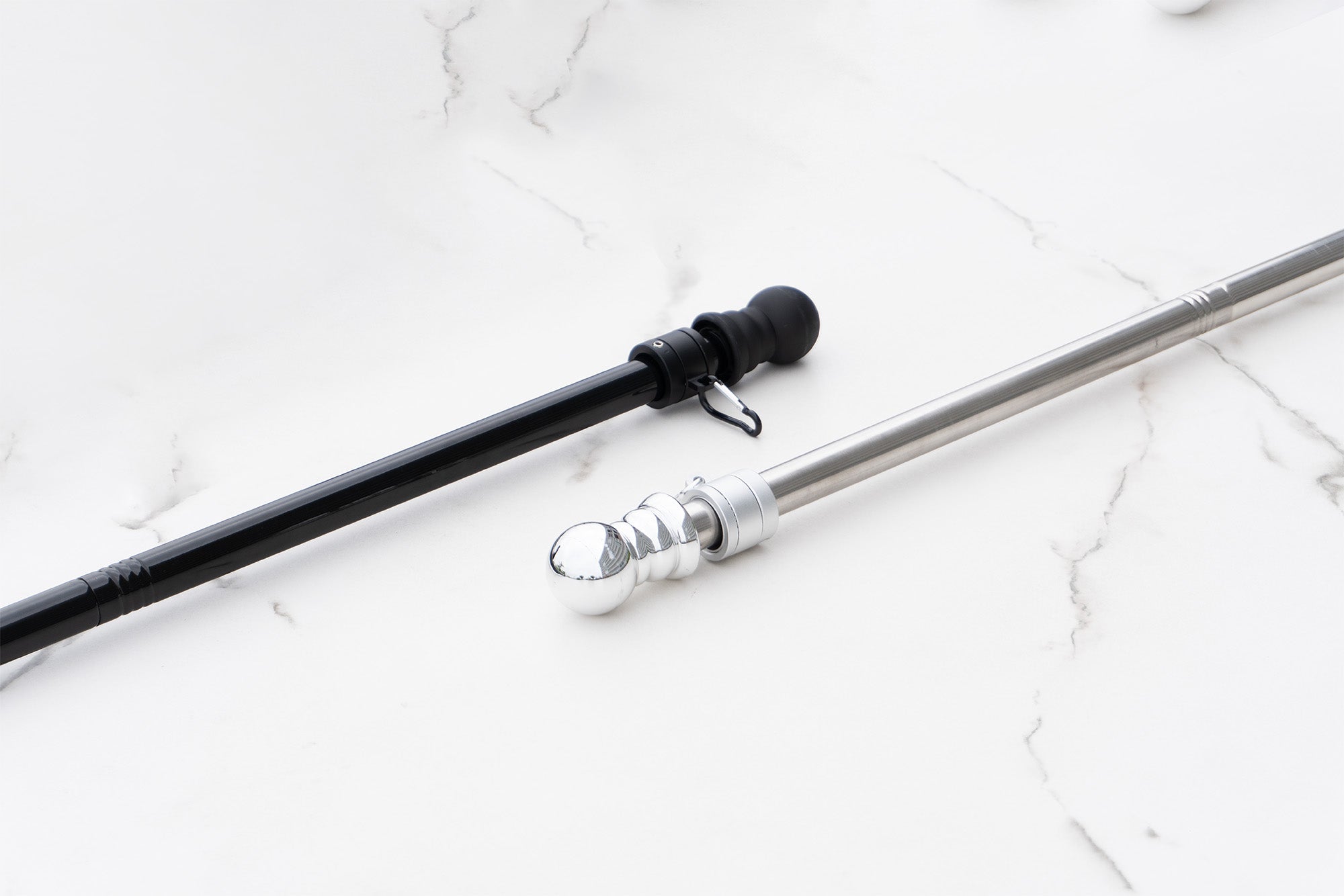 Two curtain rods on a white marble surface: a black rod with a cylindrical end and a silver rod with a spherical, ridged end, both expertly crafted by the manufacturer.