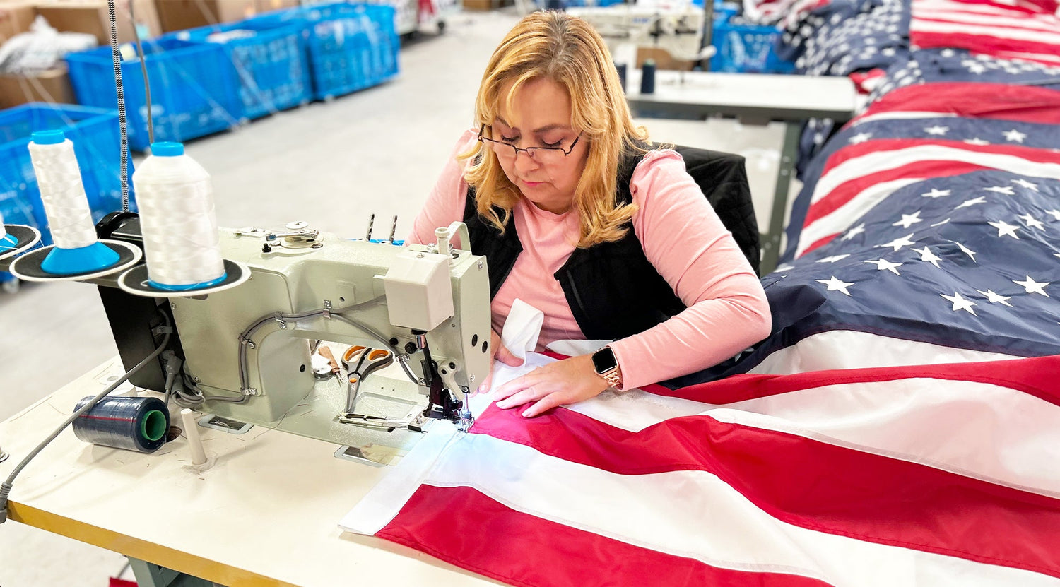 A woman works with a sewing machine to craft a long-lasting American flag in a workshop, proudly displaying the "Made in USA" label, with blue storage bins in the background.