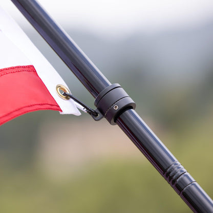 A close-up of The FlagStars' AMERICAN FLAG SET - COMPLETE BUNDLE featuring a black pole with adjustable aluminum and tangle-free spinners. The slightly blurred background accentuates the vibrant red and white flag, which is securely attached with a metal clip.