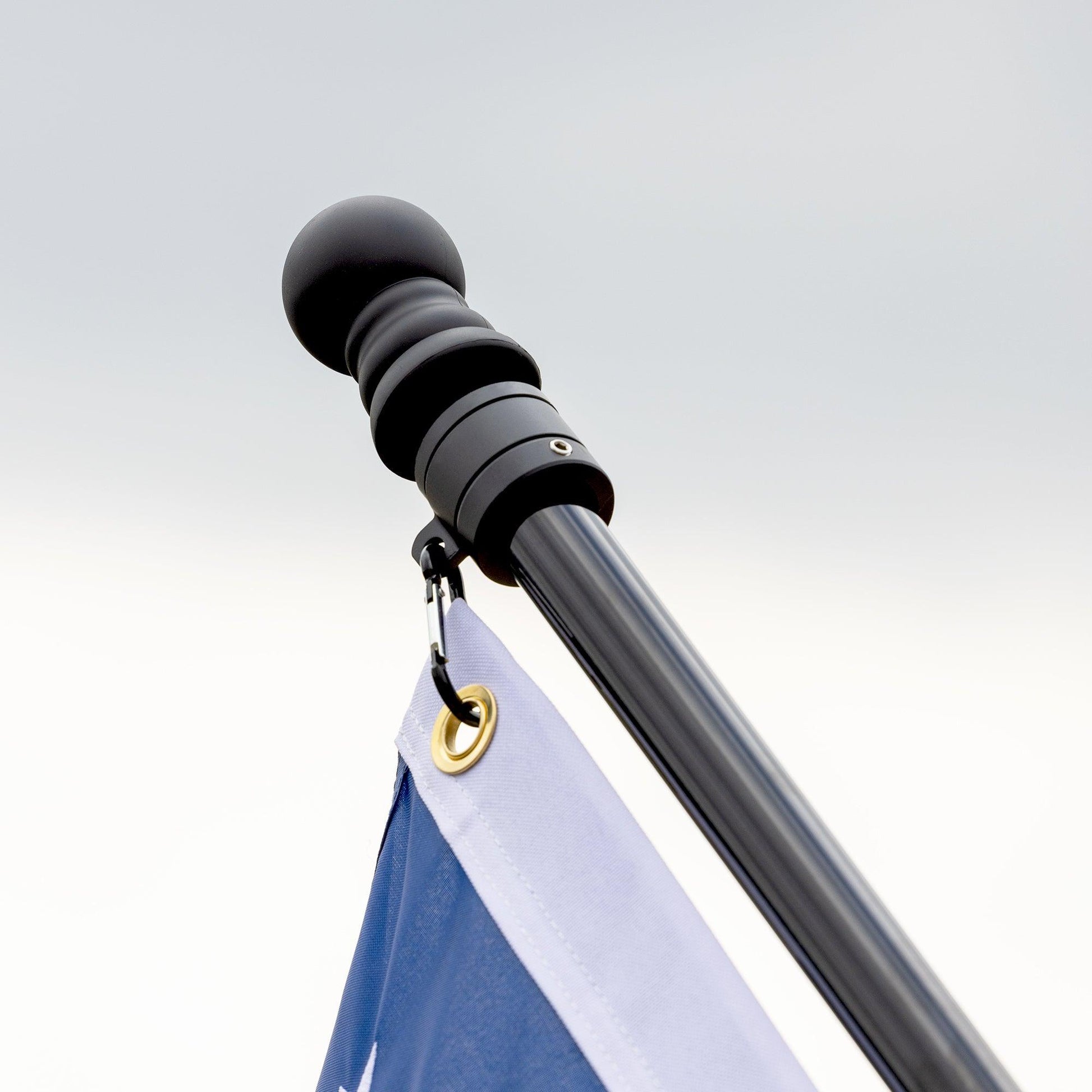 Close-up of the top of the Flag Stars Inc 5-Section 6FT Stainless Steel Flagpole, featuring a ball finial, tangle-free aluminum spinners, and a flag with a gold grommet in blue and white against a cloudy sky.