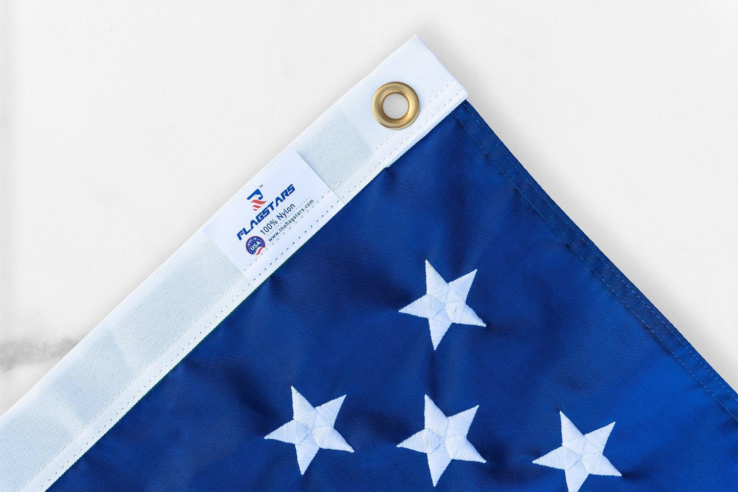 Close-up of a corner of an American flag with white stars on blue fabric, featuring a grommet in the top left and a manufacturer's tag on the white edge, proudly made in the USA. Product Name: 2' X 3' AMERICAN FLAG by Brand Name: The FlagStars.