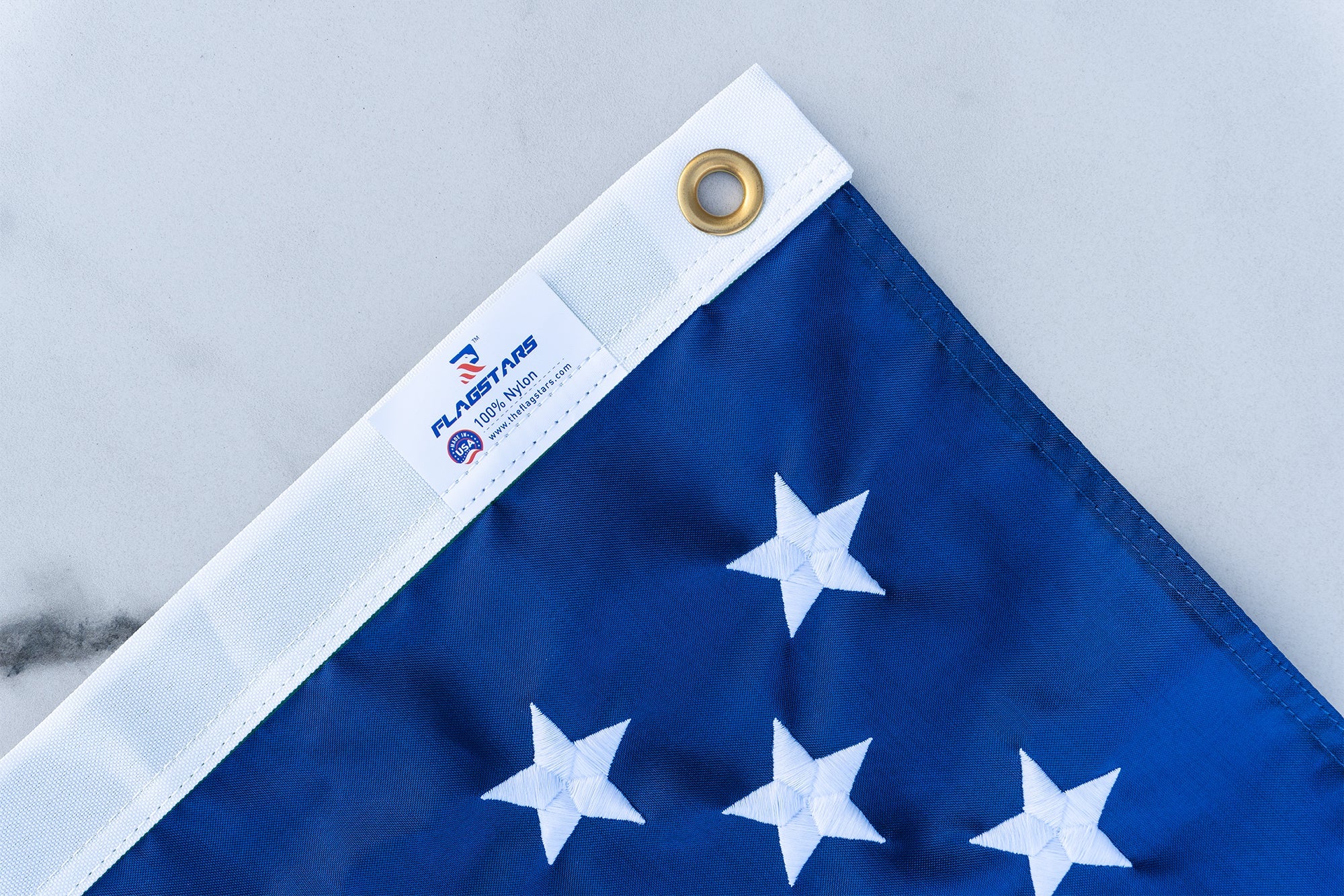 Close-up of a corner of a folded American flag, showing the blue field with white stars, brass grommet, and the label "FlagStars, made in USA," set against a light gray background.