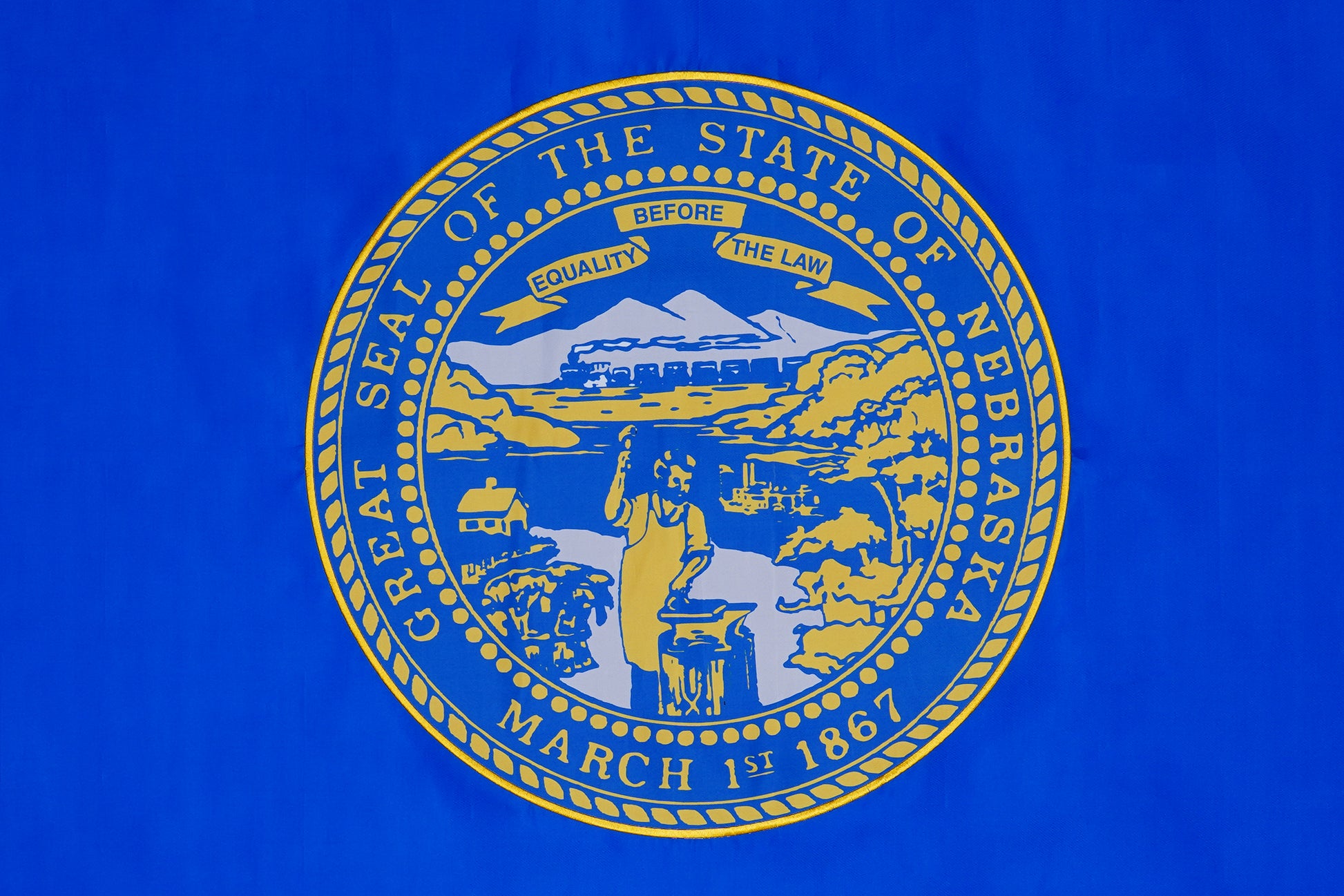 The Nebraska State Flag by Flag Stars Inc features the state seal with a blacksmith, cabins, train, and mountains on blue with detailed embroidery and "Great Seal of the State of Nebraska March 1st 1867.” 100% Made in USA. Suitable for outdoor use.