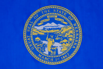 The Nebraska State Flag by Flag Stars Inc features the state seal with a blacksmith, cabins, train, and mountains on blue with detailed embroidery and "Great Seal of the State of Nebraska March 1st 1867.” 100% Made in USA. Suitable for outdoor use.
