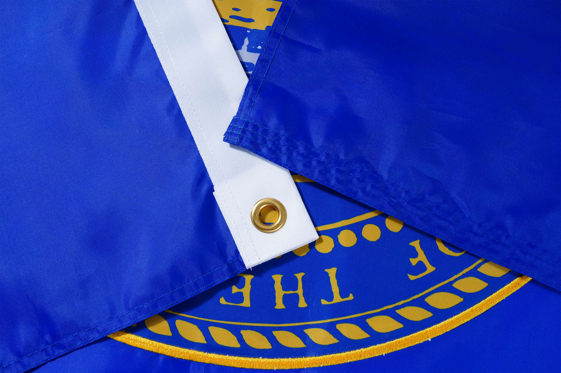 Close-up of two overlapping Nebraska State Flags by Flag Stars Inc, showing blue fabric, brass grommet, and partial gold emblem with raised embroidery—ideal for outdoor use. 100% Made in USA.