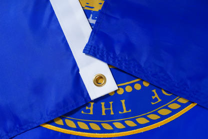 Close-up of two overlapping Nebraska State Flags by Flag Stars Inc, showing blue fabric, brass grommet, and partial gold emblem with raised embroidery—ideal for outdoor use. 100% Made in USA.