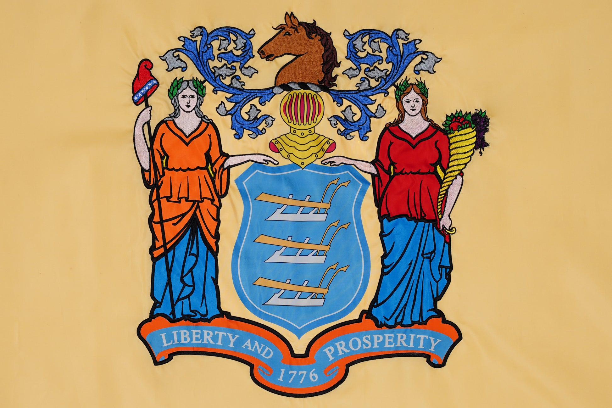 Flag Stars Inc’s New Jersey State Flag - 100% Made in USA features a blue shield with three plows, two women symbols, a horse's head crest, and "Liberty and Prosperity 1776" ribbon; crafted for outdoor durability.