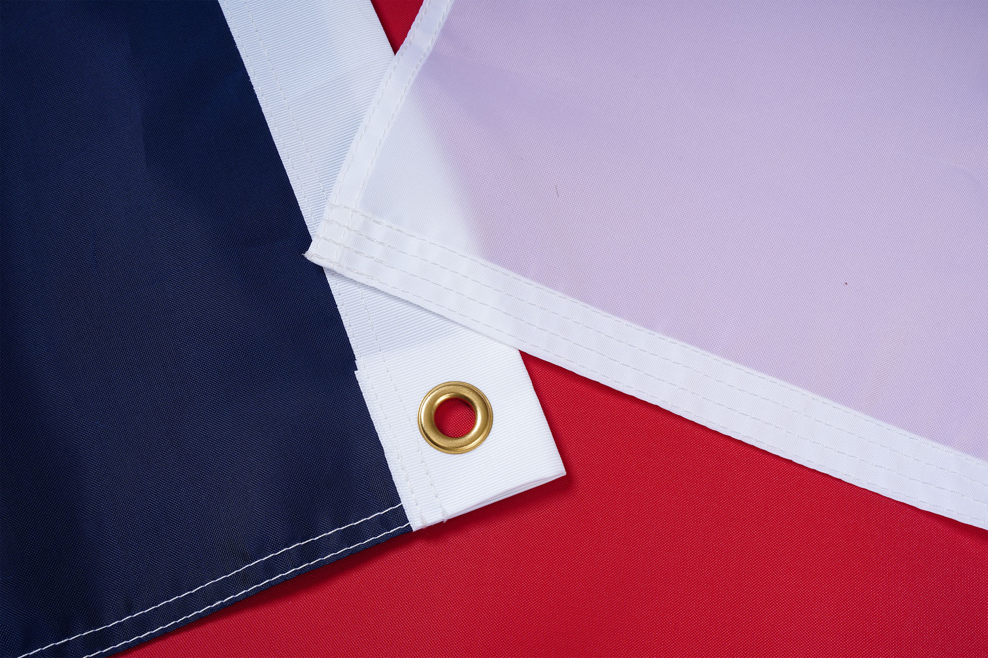 Close-up of two nylon North Carolina State Flag corners with metal grommets by Flag Stars Inc—one dark blue, one white—on a red surface. 100% Made in USA and outdoor ready.
