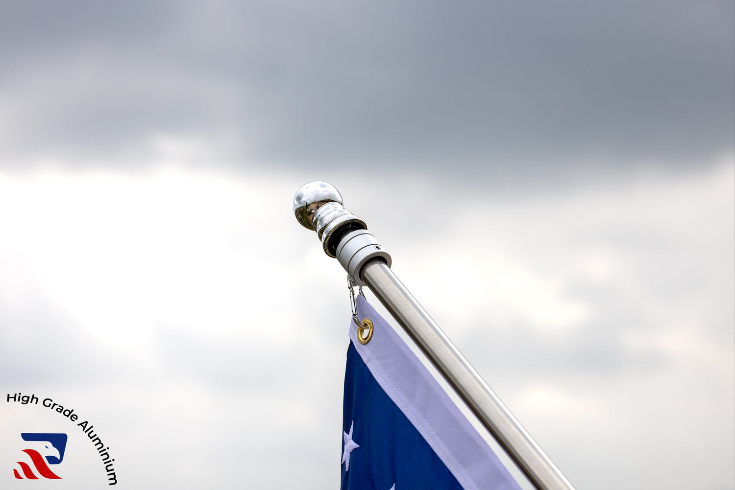 A close-up of a flagpole with a blue flag featuring white stars against a cloudy sky. There is a logo at the bottom left with the text "High Grade Aluminium, Made in USA.