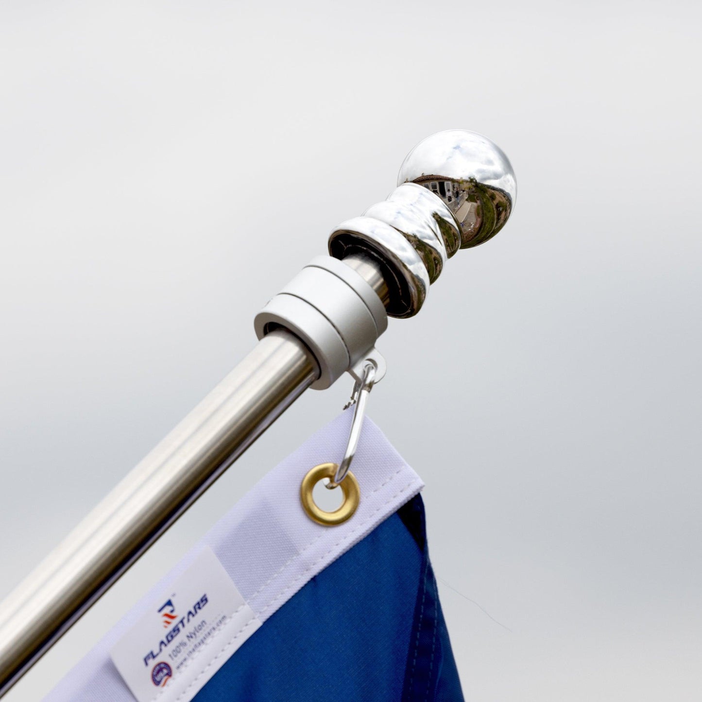 Close-up of the adjustable aluminum flagpole from the AMERICAN FLAG SET - COMPLETE BUNDLE with a blue and white flag attached. The flagpole, branded by The FlagStars, features a shiny metallic finial at the top and is equipped with tangle-free spinners. A small label on the flag reads "The FlagStars.