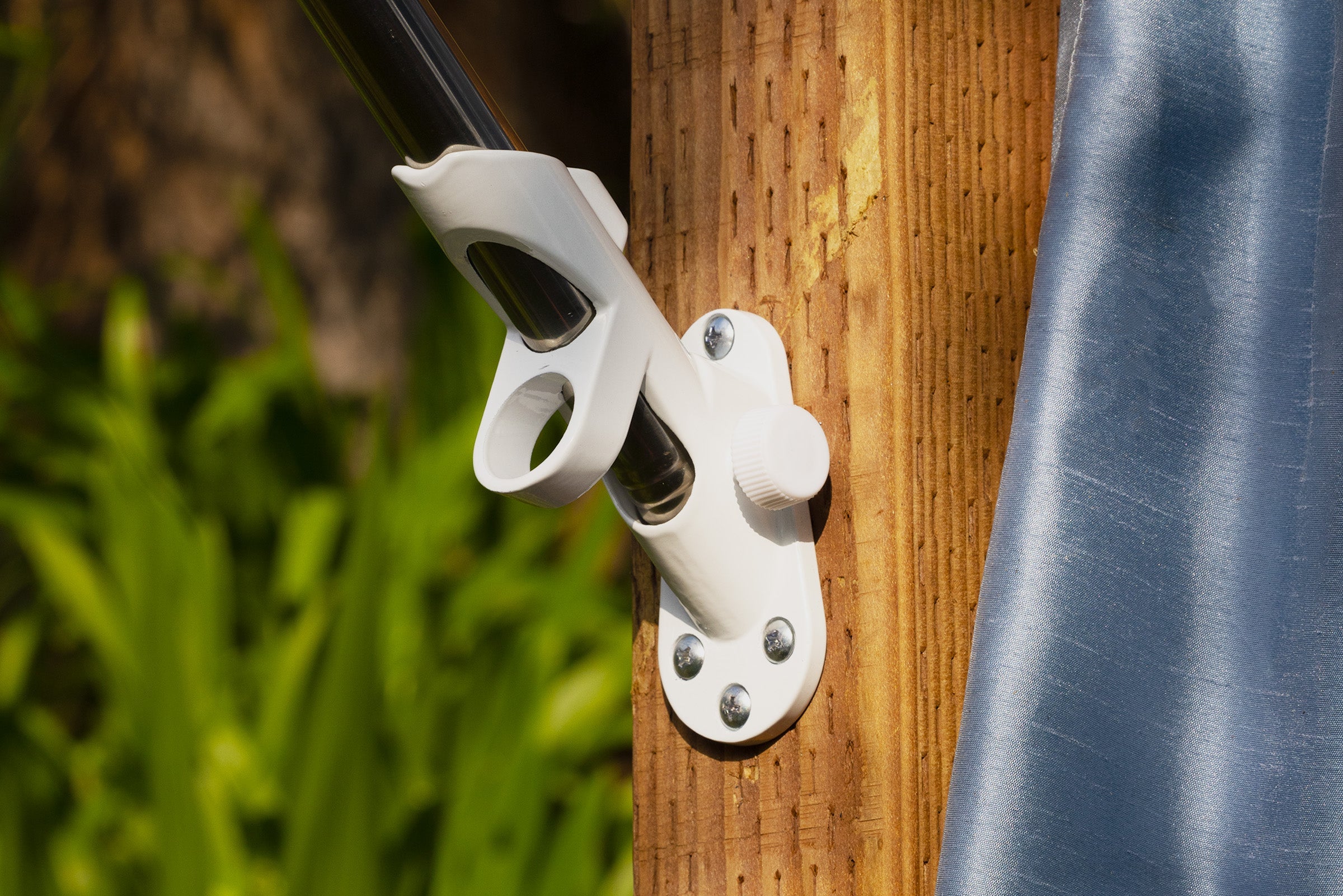A pre-assembled white flag pole holder is mounted on a wooden post with screws, holding a long-lasting metal pole against a background of greenery and a blue fabric, perfect for displaying your American Flag.
